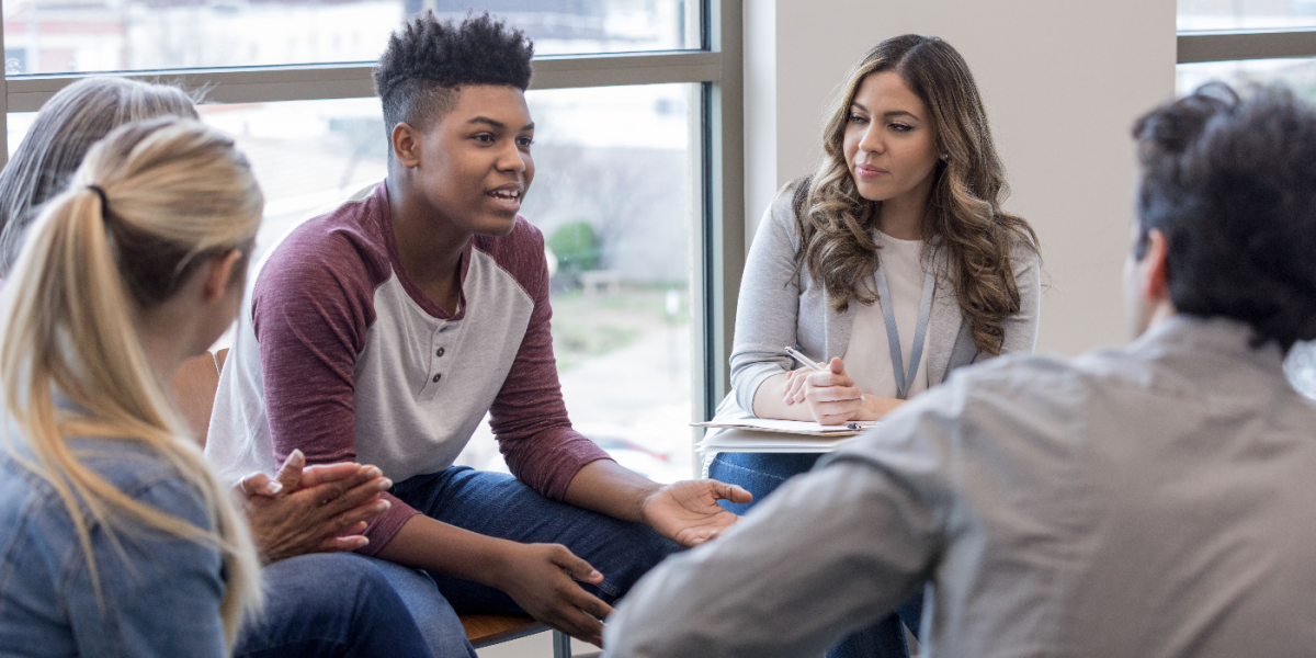 A young man shares with a larger group while a woman takes notes. 